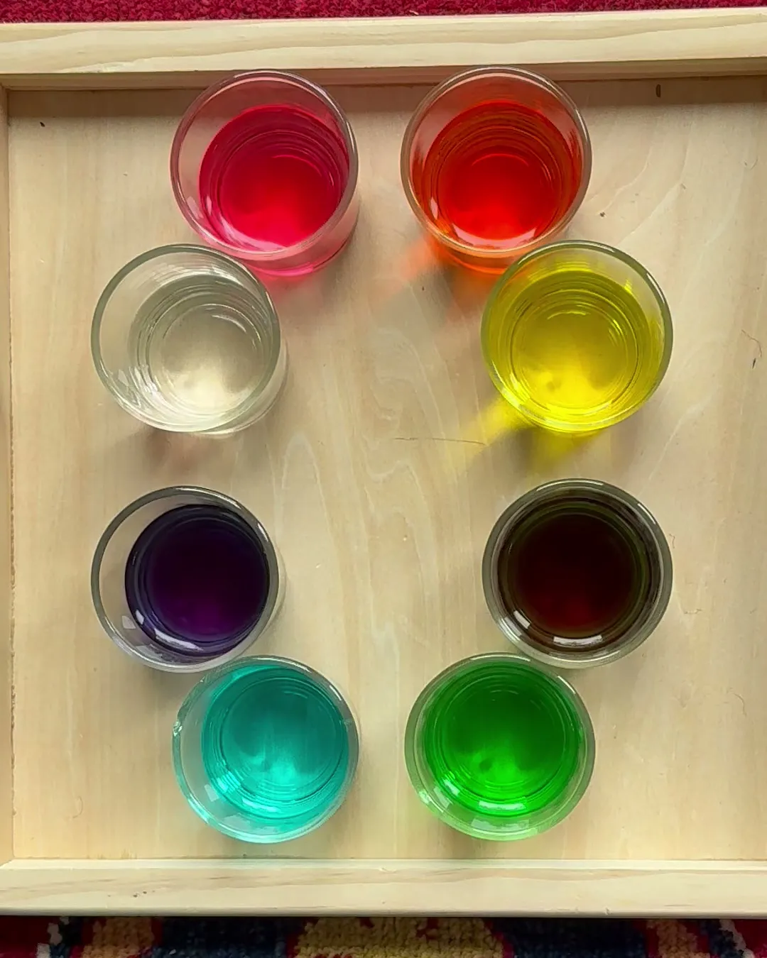 Overhead view of eight small glasses of coloured water arranged in a ring on a wooden tray: red, orange, yellow, brown, green, teal, purple and one left clear.